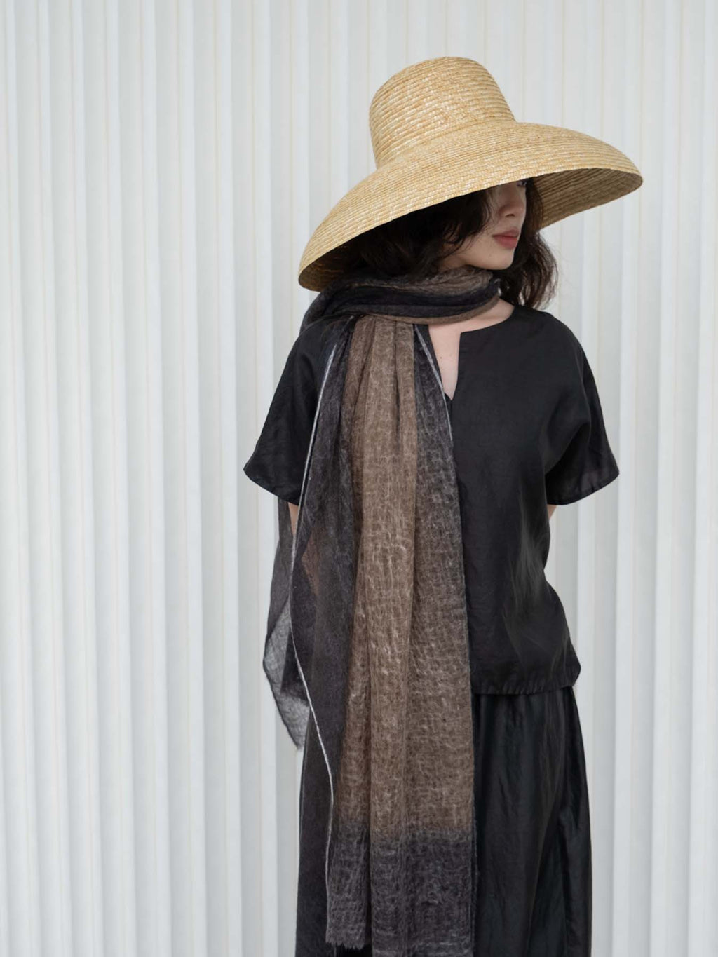 A woman wearing a wide-brimmed straw hat, a black short-sleeve dress, and a dark brown and black sheer scarf, standing against a white ribbed wall.