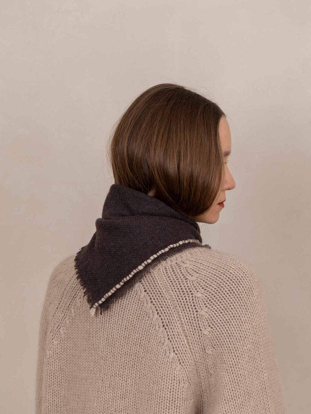 A woman with a short brown bob haircut is shown from the back and side profile, wearing a dark charcoal knit scarf with a fringed edge over a beige cable-knit sweater against a plain beige background.