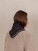 A woman with a short brown bob haircut is shown from the back and side profile, wearing a dark charcoal knit scarf with a fringed edge over a beige cable-knit sweater against a plain beige background.