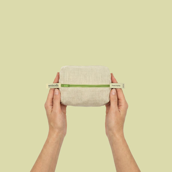 A person's hands holding a small rectangular beige linen pouch with a green band and white end-tags against a solid pale yellow-green background.
