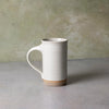 A tall, cylindrical ceramic mug with a curved handle, featuring a glossy off-white glaze and a natural, unglazed tan clay base, set against a textured light gray and beige background.