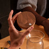 A hand holds a round wooden lid with a small clear droplet on it above a glass carafe on a wooden table, with another person's arm visible in the background.