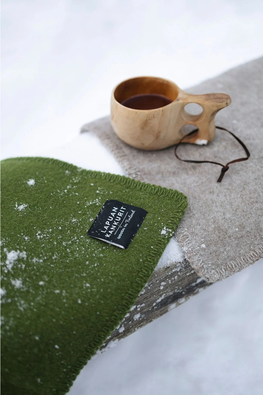 Two textured woolen seat pad, one olive green with a black 'LAVUAN KANJUIT' label and one light gray, styled outdoors in the snow with a small wooden cup and string.