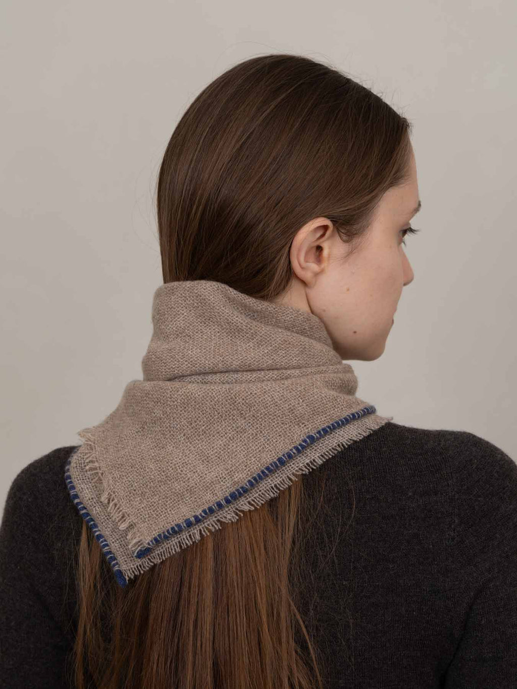 A woman shown from the back and side profile wearing a beige textured scarf with blue fringe trim around the neck.