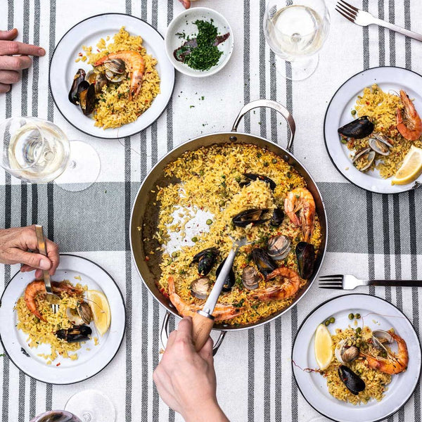 Overhead view of a seafood paella spread on a striped tablecloth, with a large pan of yellow rice, shrimp, mussels, and fish in the center and several white plates being served by visible hands.