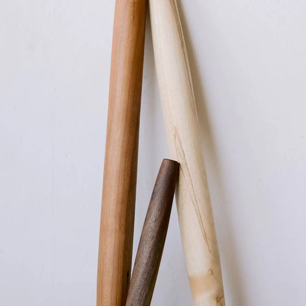 Three wooden rolling pins in natural light brown, cream, and dark brown tones, arranged vertically against a plain off-white background.