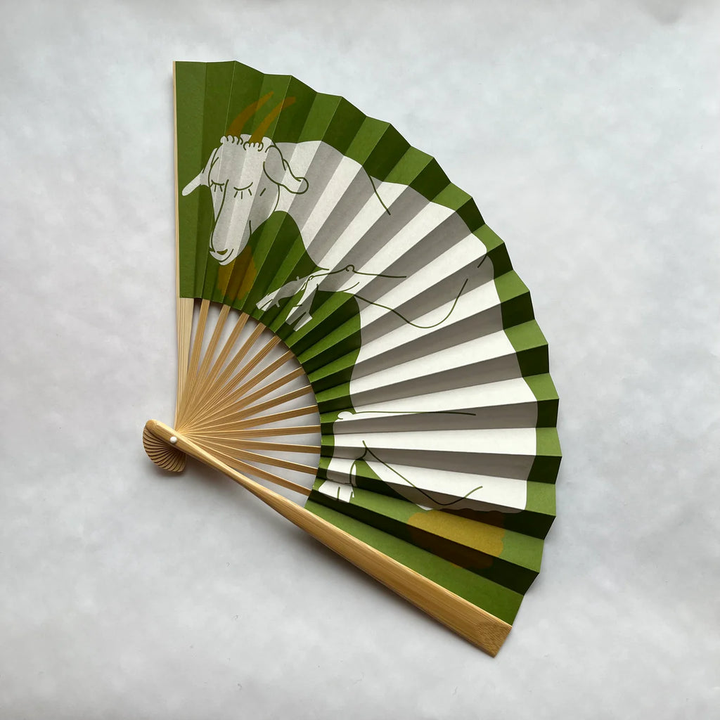 Handheld fan with green and brown and white cow design featuring a goat on a light gray background