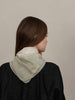 A woman shown from the back and side profile wearing a light beige knit hood or cowl with a scalloped edge over a black textured top, set against a plain beige studio background.