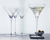 Four clear martini glasses on a gray surface against a white background, with one glass filled with a pale drink and a small dish of olives beside it.