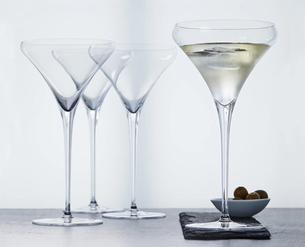 Four clear martini glasses on a gray surface against a white background, with one glass filled with a pale drink and a small dish of olives beside it.