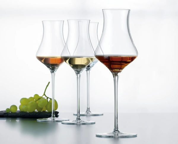 Four clear stemmed wine glasses arranged in a row on a light gray surface against a white background, with green grapes on a dark plate at the left and wine in the front two glasses.