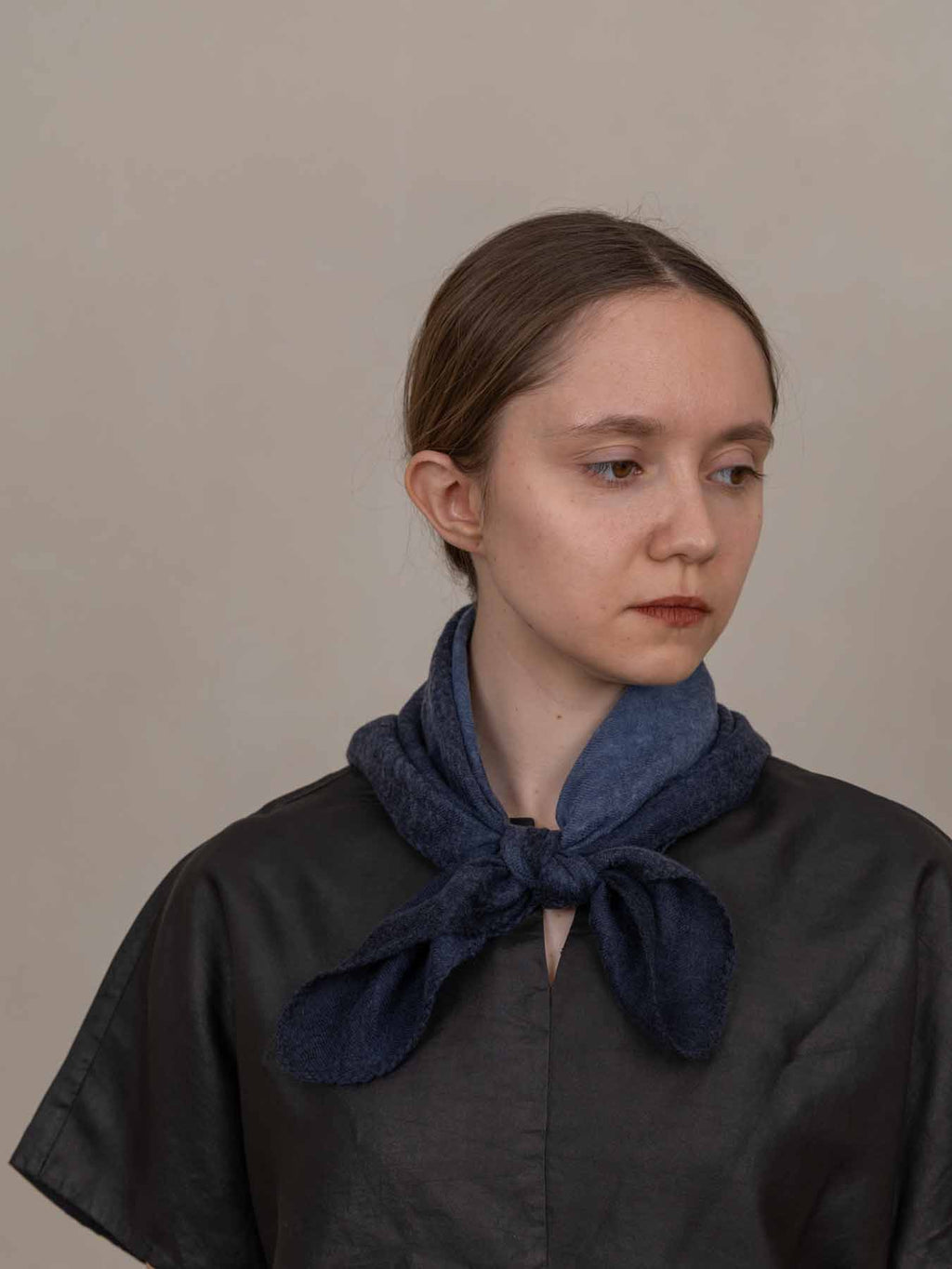 A woman wearing a dark charcoal gray top and a dark blue textured scarf tied around her neck, photographed against a plain beige background.