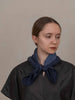 A woman wearing a dark charcoal gray top and a dark blue textured scarf tied around her neck, photographed against a plain beige background.