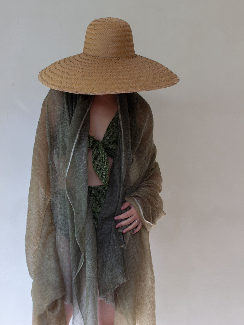 A person wearing a large woven straw sun hat, a sheer taupe-beige shawl draped over an olive green bikini, standing against a plain off-white background.