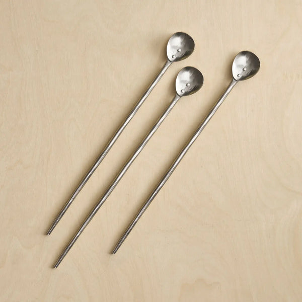 Three long metal stirring spoons with hammered oval heads, arranged diagonally on a light beige wood background.