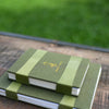 Two green books with a visible brand logo on a wooden surface with grass in the background
