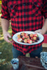 Person wearing a red and black plaid shirt holding a plate of food outdoors.