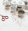 Scissors and pins on a white surface with small containers in the background.